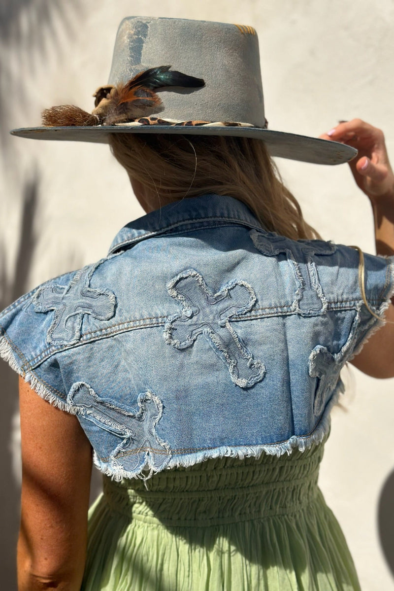 Person wearing a denim vest with floral patterns and a wide-brimmed hat with feathers.